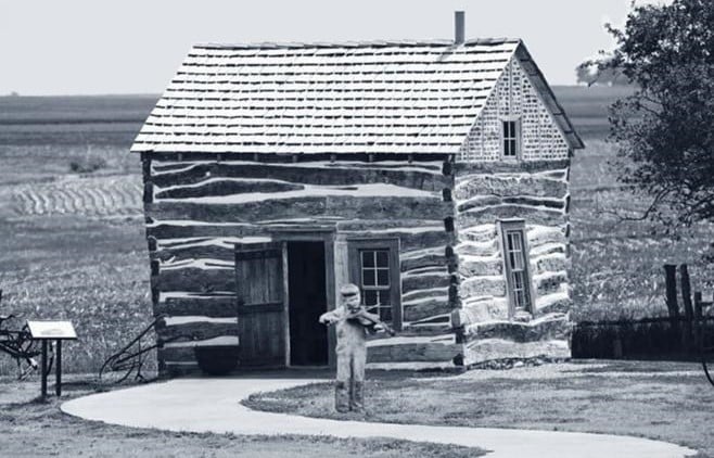 Black and white photo of a boy playing a fiddle in front of a small log cabin