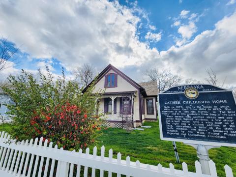Foreground is a pink-blooming bush, a white picket fence, and a historic marker sign titled Cather Childhood Home. Background is a small frame house with a front porch.
