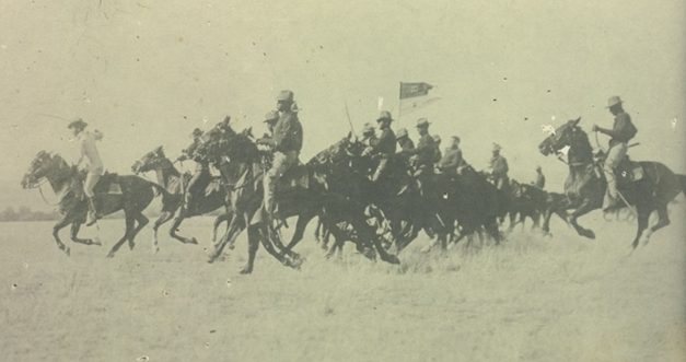 sepia photo of a group of about a dozen horses ridden by soldiers with a military flag visible above the group