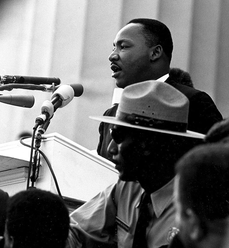 Martin Luther King Jr. speaks into a microphone. A fluted building column is visible behind him, and a uniformed Park Ranger stands near him.