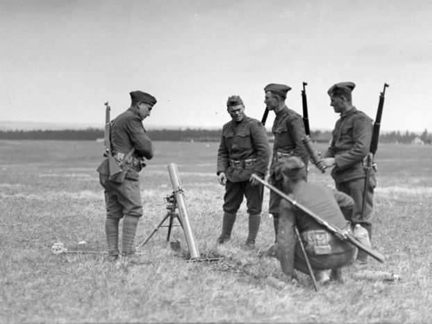 Black and white photo of five World War I soldiers, all with rifles slung across their backs, gathered around a tube-shaped piece of equipment on a tripod.