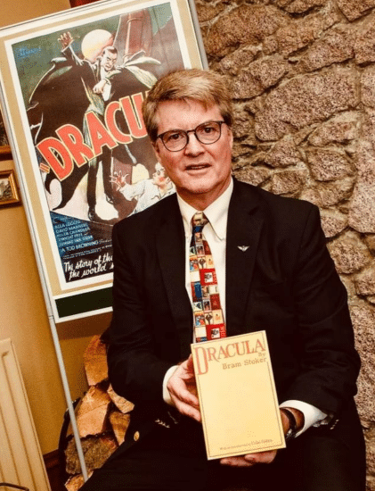 Dacre Stoker holds a copy of the novel "Dracula". A Dracula movie poster is behind him. He's wearing a tie patterned with vampire images.