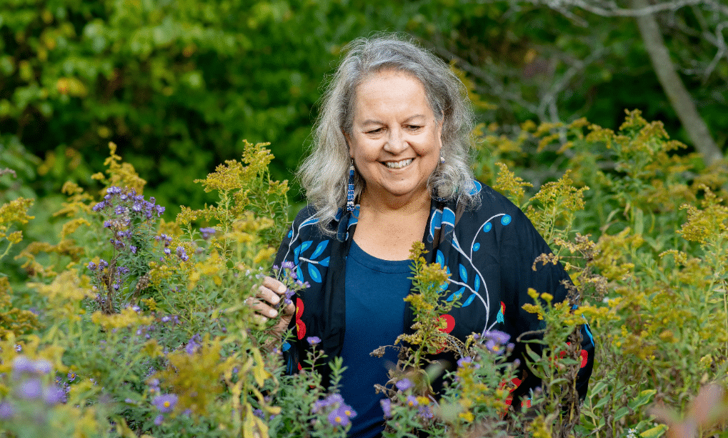 A smiling woman with gray hair stands among shoulder-high prairie flowers. She does not look into the camera. She holds one of the plants in her hand.