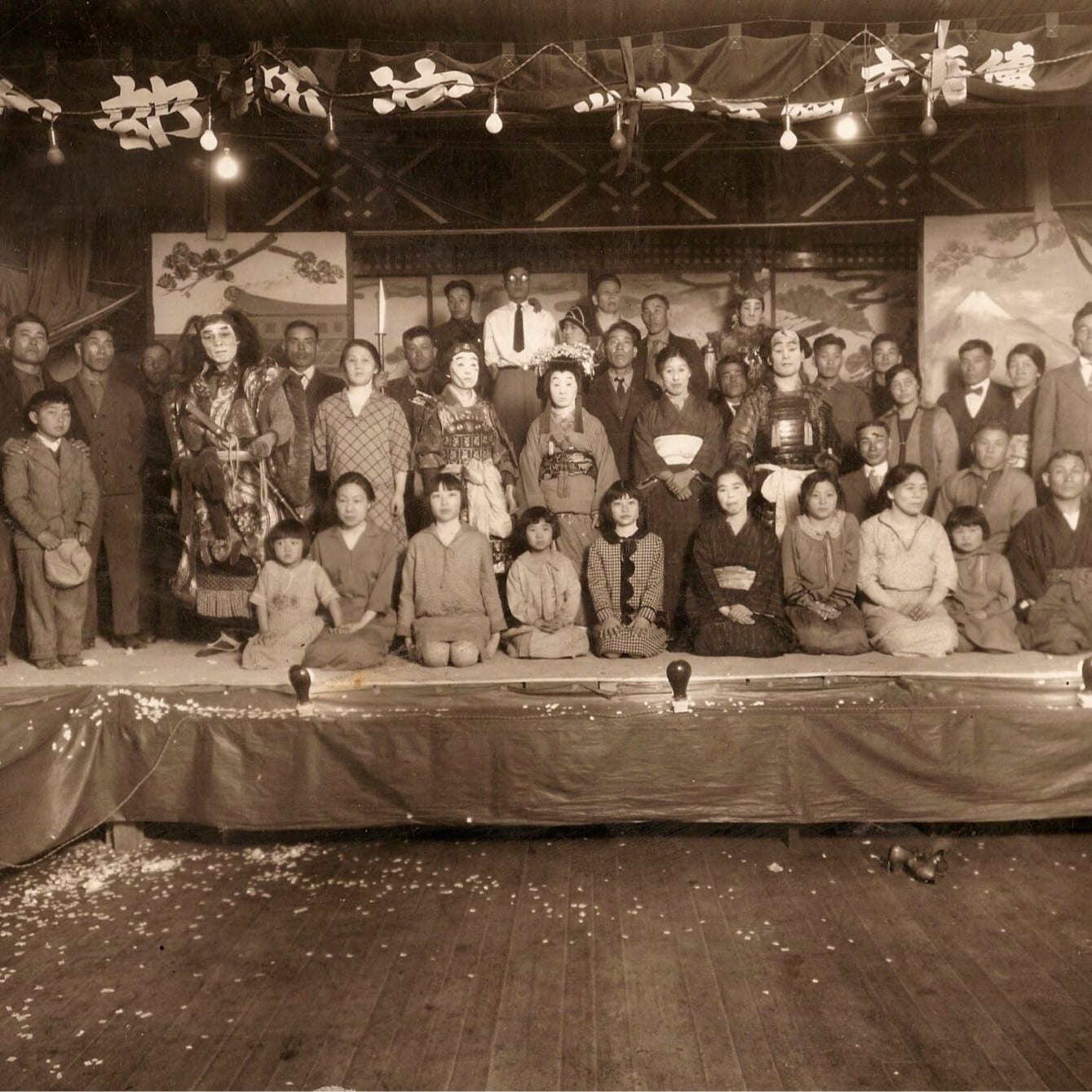 Black and white photo of rows of Japanese people on stage, some in theater costumes and masks.