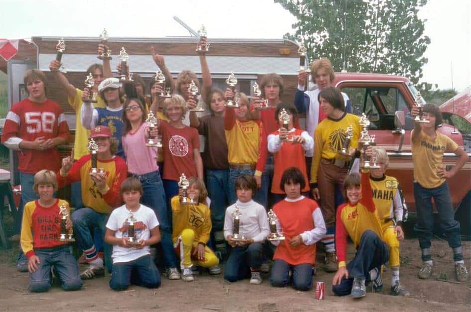 A group of young men / boys in what look like 1970s or early 1980s jeans/tshirts and hairstyles hold up trophies that feature a bicyclist on top. A pickup truck with a camper topper is in the background.