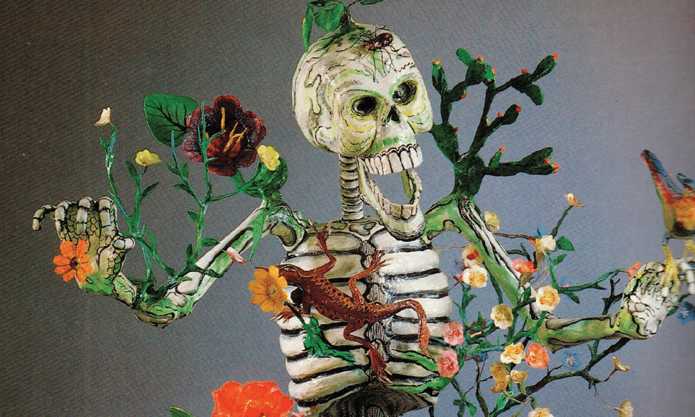 Photo of a skeleton sculpture. Various sculpted plants grow from the skeleton, including flowers and a cactus. A lizard its on the skeleton's ribcage, and a spider sits on its head.