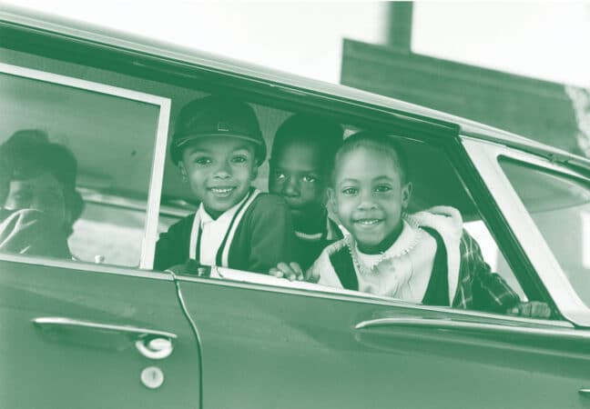 A grayscale photo produced in green that shows three children looking out the open back seat window of a car. An adult woman is visible in the front seat in the background.