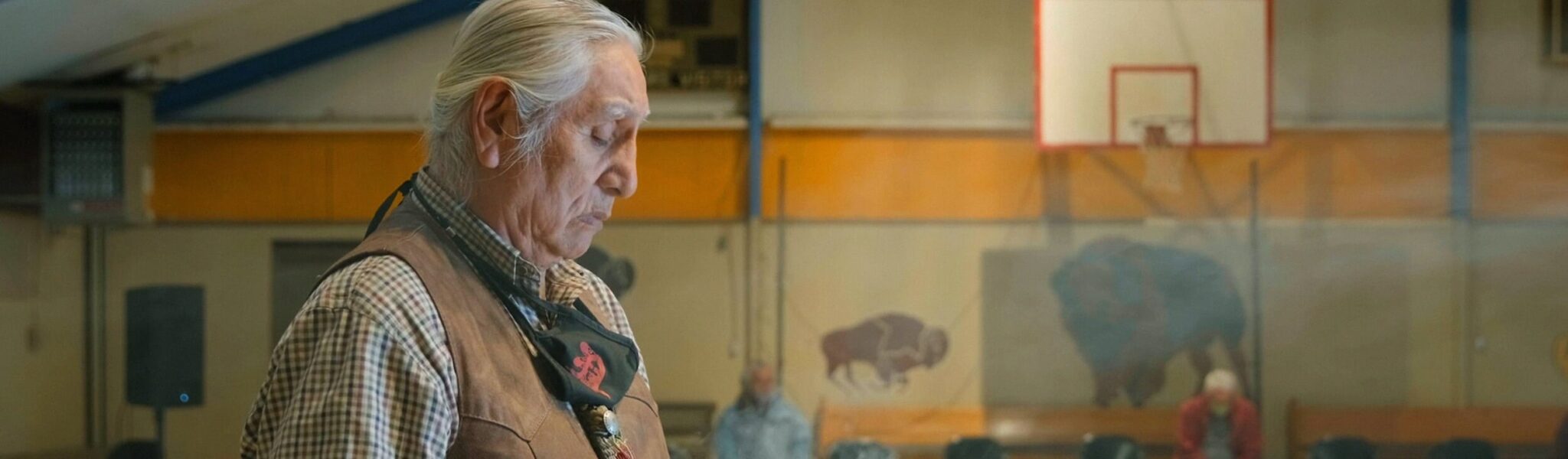 An older man in a checkered shirt and leather vest looks down. Behind him is the wall of a gymnasium with a basketball net. There are bison painted on the wall.