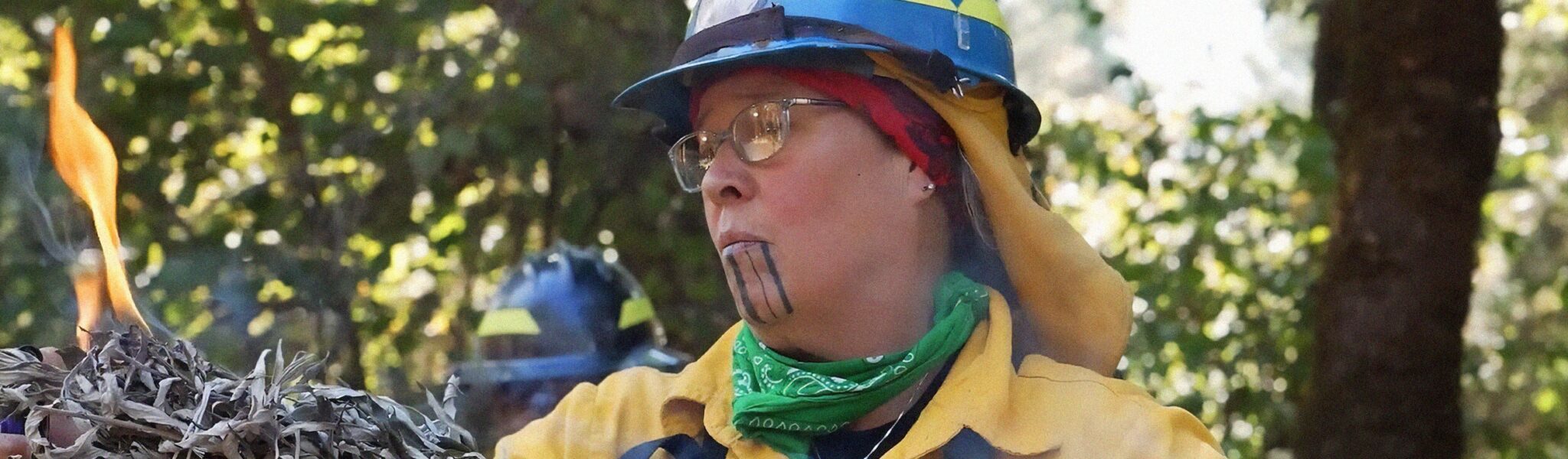A woman wearing fire gear who has a tattoo on her chin looks into the flame of burning sage. Green tree leaves are visible behind her.