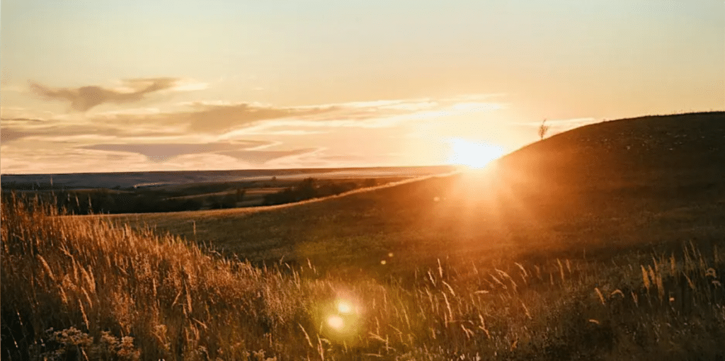 Landscape photo of rolling prairie grass hills with the sun on the horizon.