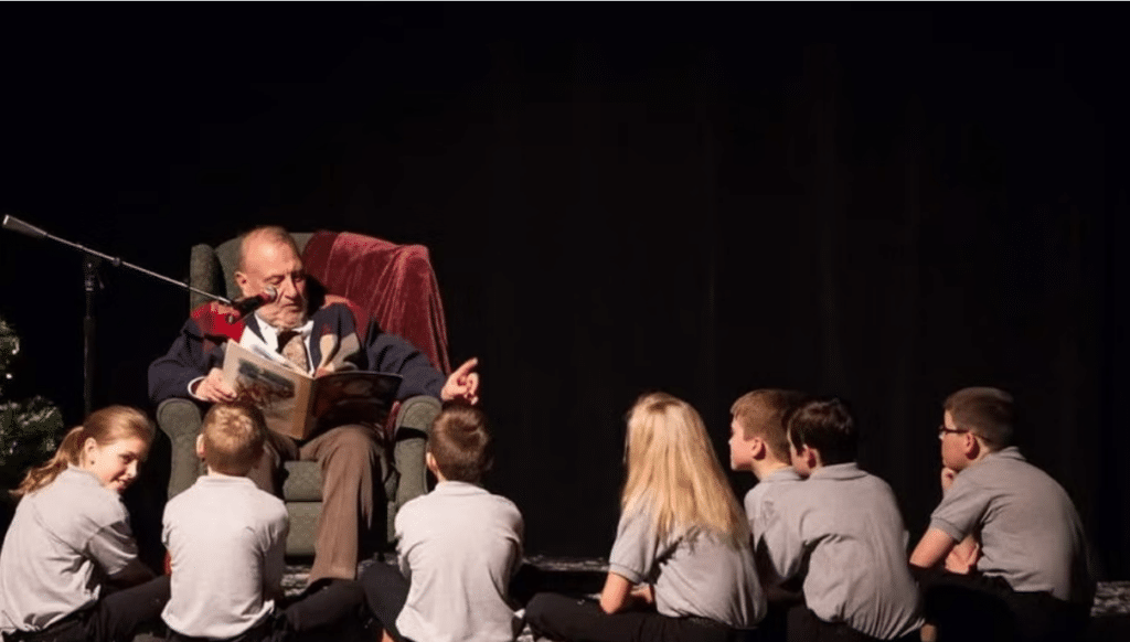 Chuck Peek sits in a chair with a book in his lap and a microphone in front of him reading to a group of children sitting on the floor.