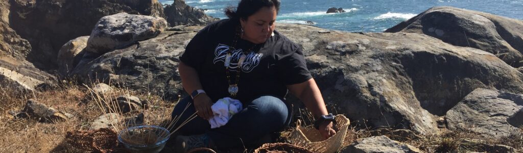 Woman sitting on the ground in a rocky coastal area with baskets and basket-making materials in front of her.