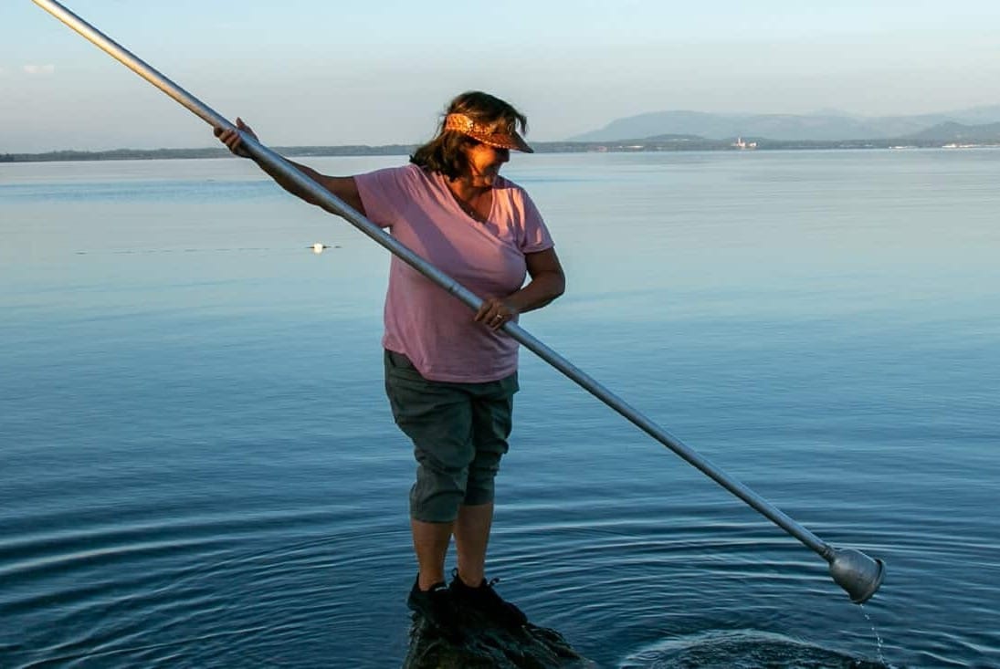 Woman standing on a rock in a large body of water, holding a large metal pole and dipping it towards the water