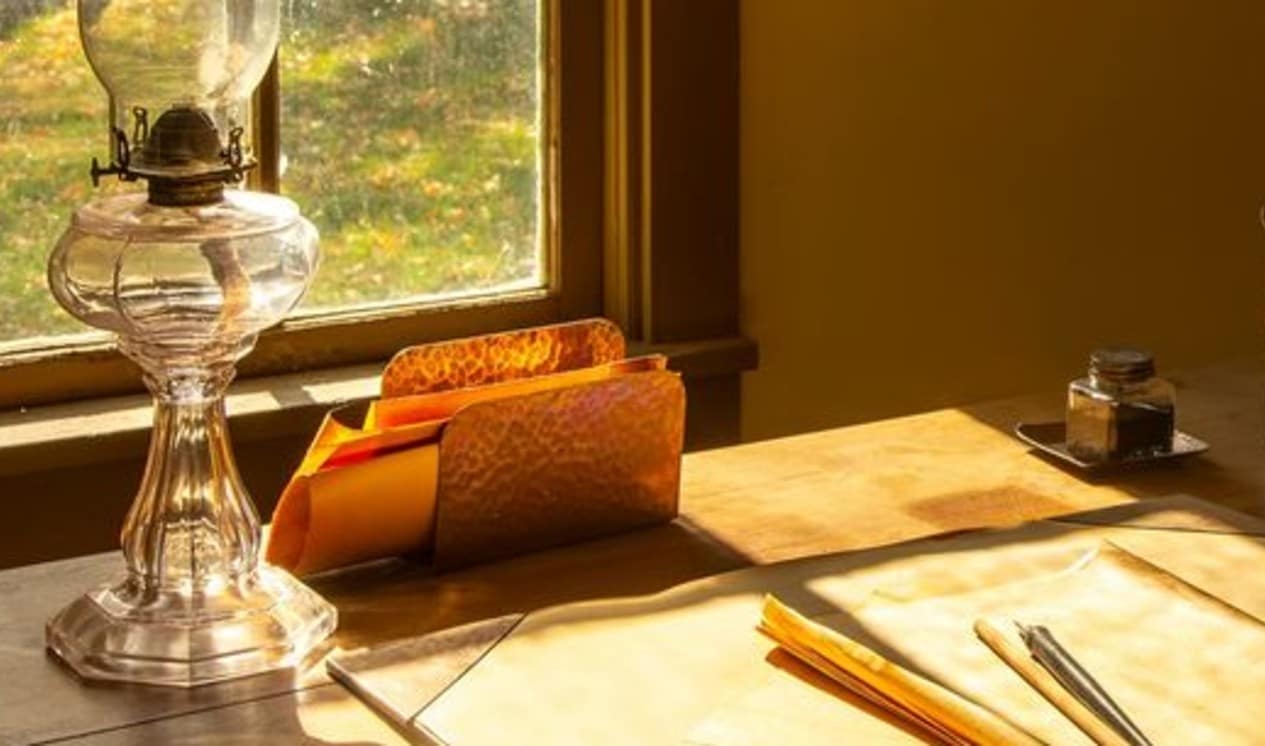 Papers and an oil lamp on a desk in front of a window.