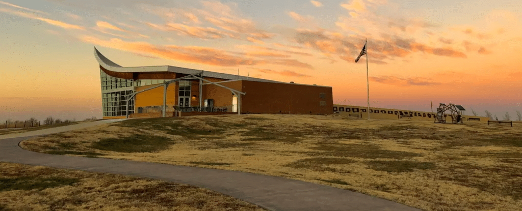 The plow shaped visitor center is in the foreground. A pale pink sky can be seen behind it as the sunsets. Sunset at the Homestead Heritage Center. NPS Photo/H Hendricks