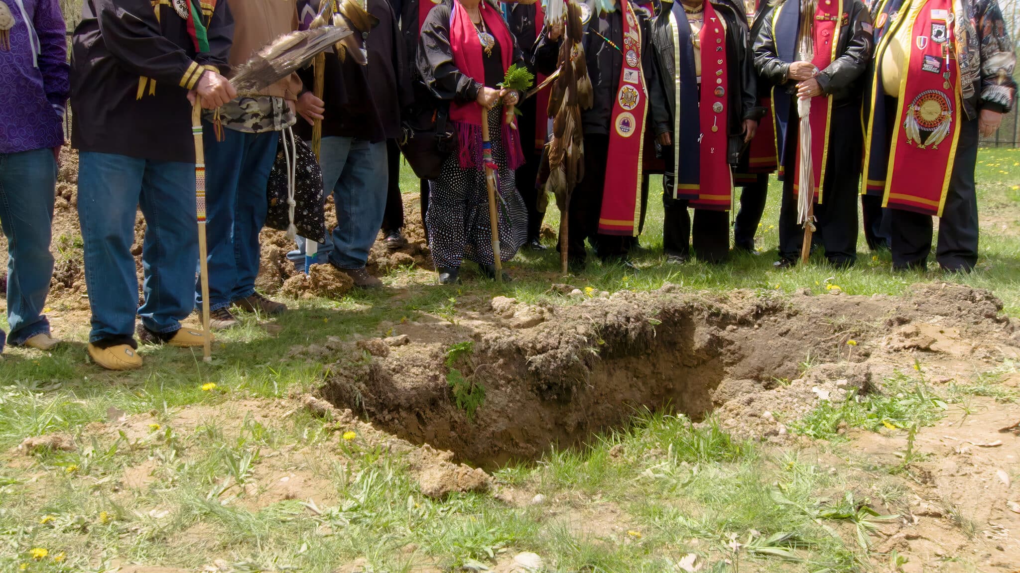 Several people visible from the wait down stand around a deep, rectangular hole dug into the earth. Some of the people hold or are wearing symbols of Native American culture.