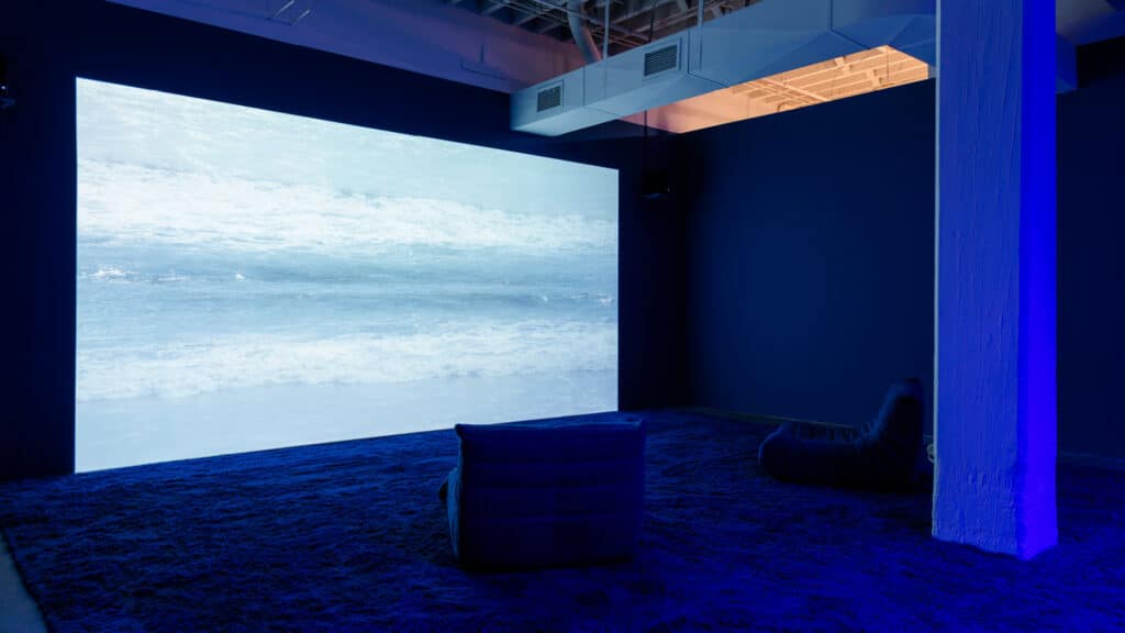 An image of small waves coming ashore is projected on a wall, and the floor appears to be the texture of rippled water. Two lounge chairs face the wall.