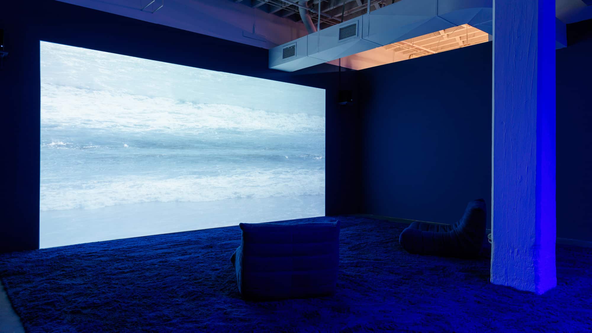 An image of small waves coming ashore is projected on a wall, and the floor appears to be the texture of rippled water. Two lounge chairs face the wall.