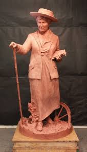 Willa Cather sculpture. She has a walking stick in one hand and a book or papers in the other. A partially buried wagon wheel is near her feet.