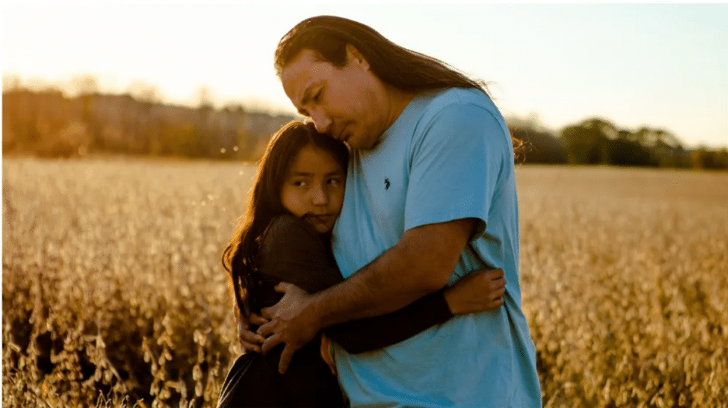 A man with long hair embraces a youth with long hair in a field backlit by the sun.