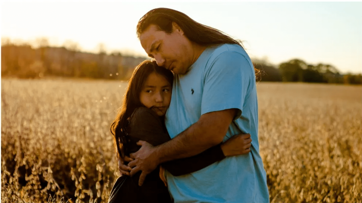 A man with long hair embraces a youth with long hair in a field backlit by the sun.